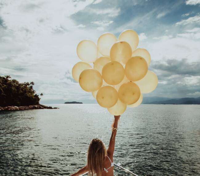 A vertical shot of a woman in a dress standing on a yacht, holding golden balloons while sailing